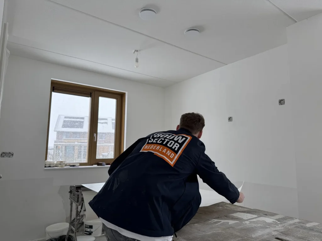 Behanger in donkere werkjas buigt over de plaktafel in een witte nieuwbouwkamer met houten raamkozijn